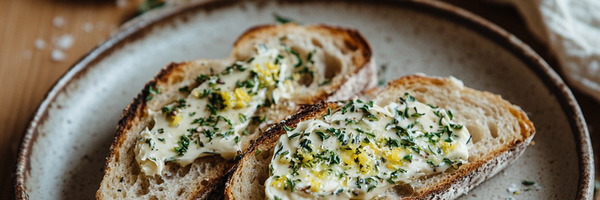 Geröstetes Sauerteigbrot mit Bärlauchbutter, bestreut mit getrockneten Kräutern und Zitronenzesten auf Keramikteller – serviert auf rustikalem Holztisch.